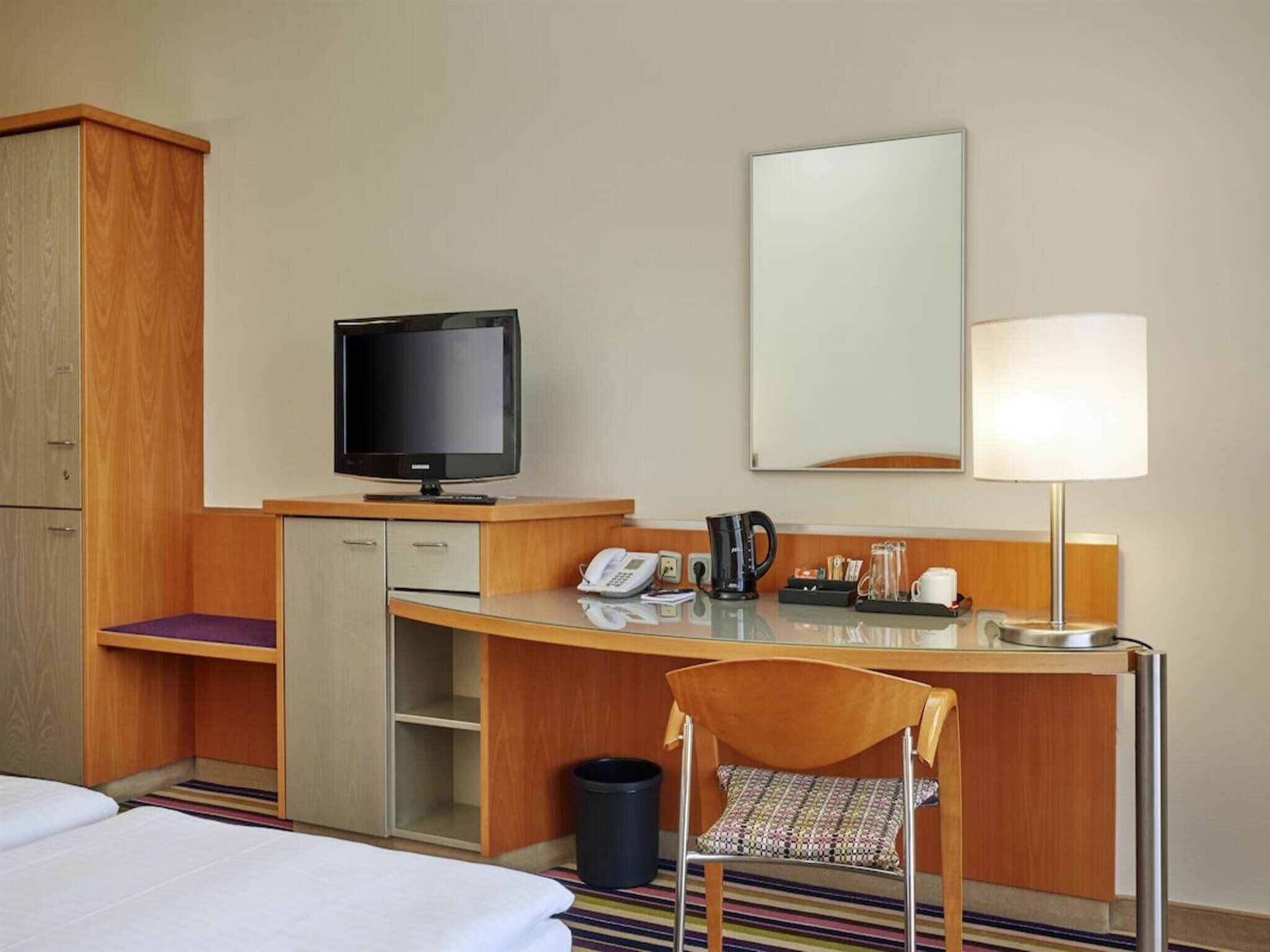 Une chambre d’hôtel comprenant un lit, un bureau avec un téléphone, une télévision, une machine à café, une chaise et une poubelle.