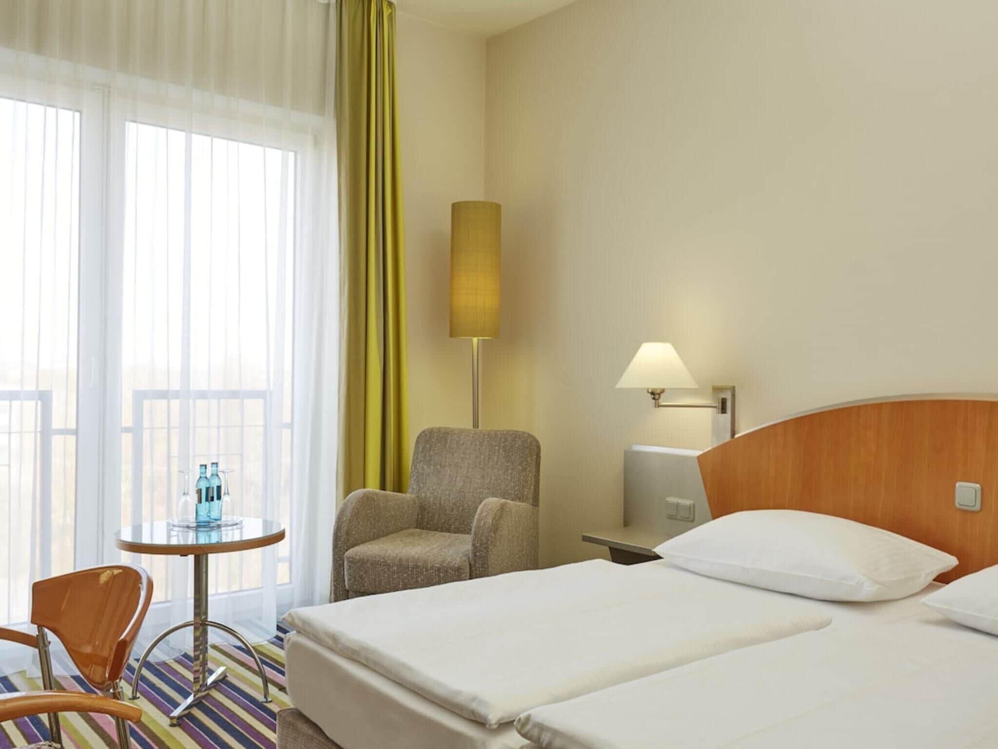 A hotel room with a bed, a chair, a small table with bottles, a lamp, and a window with curtains.