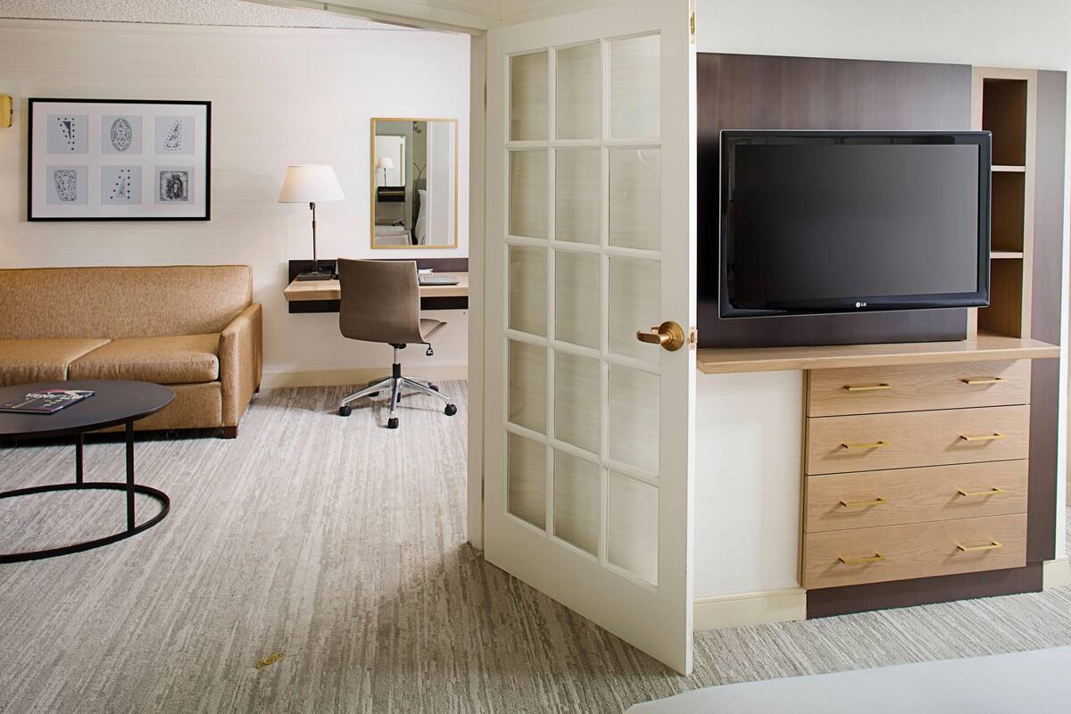 A hotel room with a sofa, a desk with a lamp, a TV on a wooden cabinet, and a door with frosted glass panels.