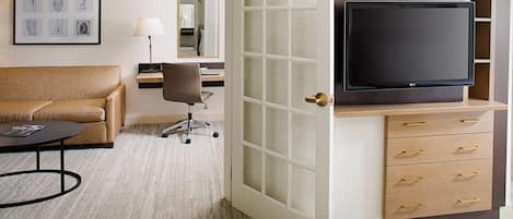 Una habitación de hotel con sofá, escritorio con lámpara, televisor sobre un mueble de madera y puerta con paneles de vidrio esmerilado.