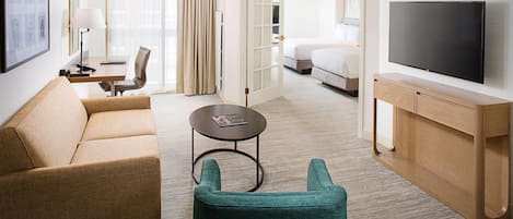Habitación de hotel con sofá, un sillón verde azulado, una mesita redonda, un televisor sobre un mueble de madera y una cama al fondo.