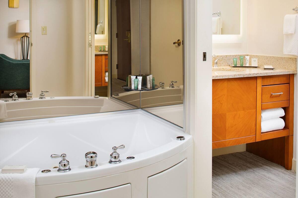 A bathroom with a large bathtub, a sink, and a mirror. There are toiletries on the sink counter and a towel rack with towels.