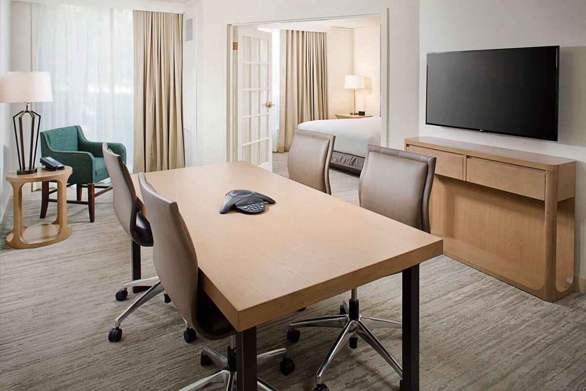 A hotel room with a large bed, a TV mounted on the wall, a desk with chairs, and a small table with a lamp.