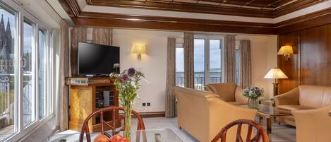 A spacious living room with a large flat-screen TV, comfortable seating, a glass coffee table, and a view of a historic building through large windows.