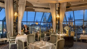 A restaurant with tables set for dining, large windows offering a city view, and a chandelier.