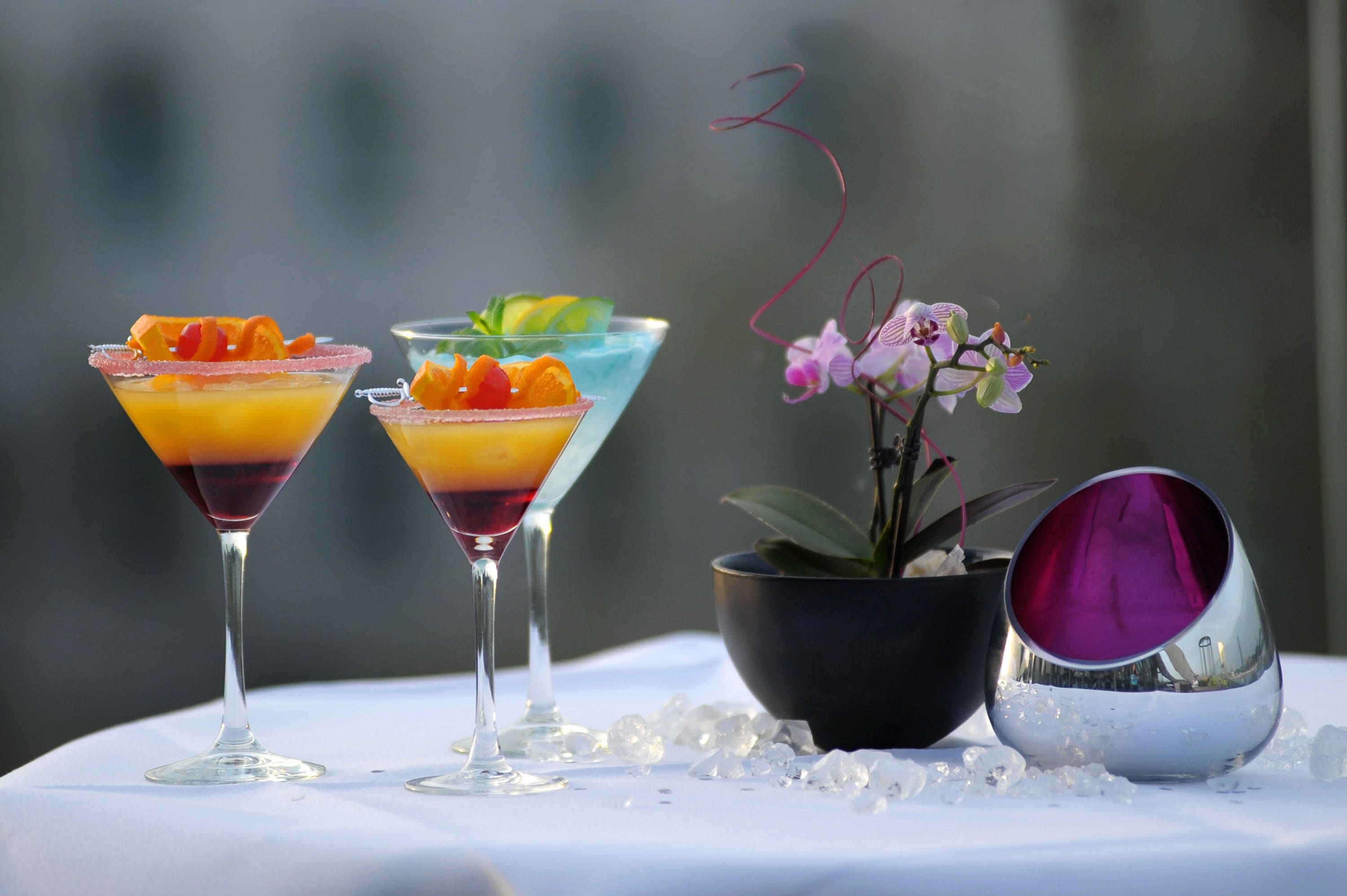 Deux verres à martini contenant des cocktails à plusieurs couches et décorés de fruits, une orchidée en pot et un bol décoratif posés sur une table avec des glaçons.