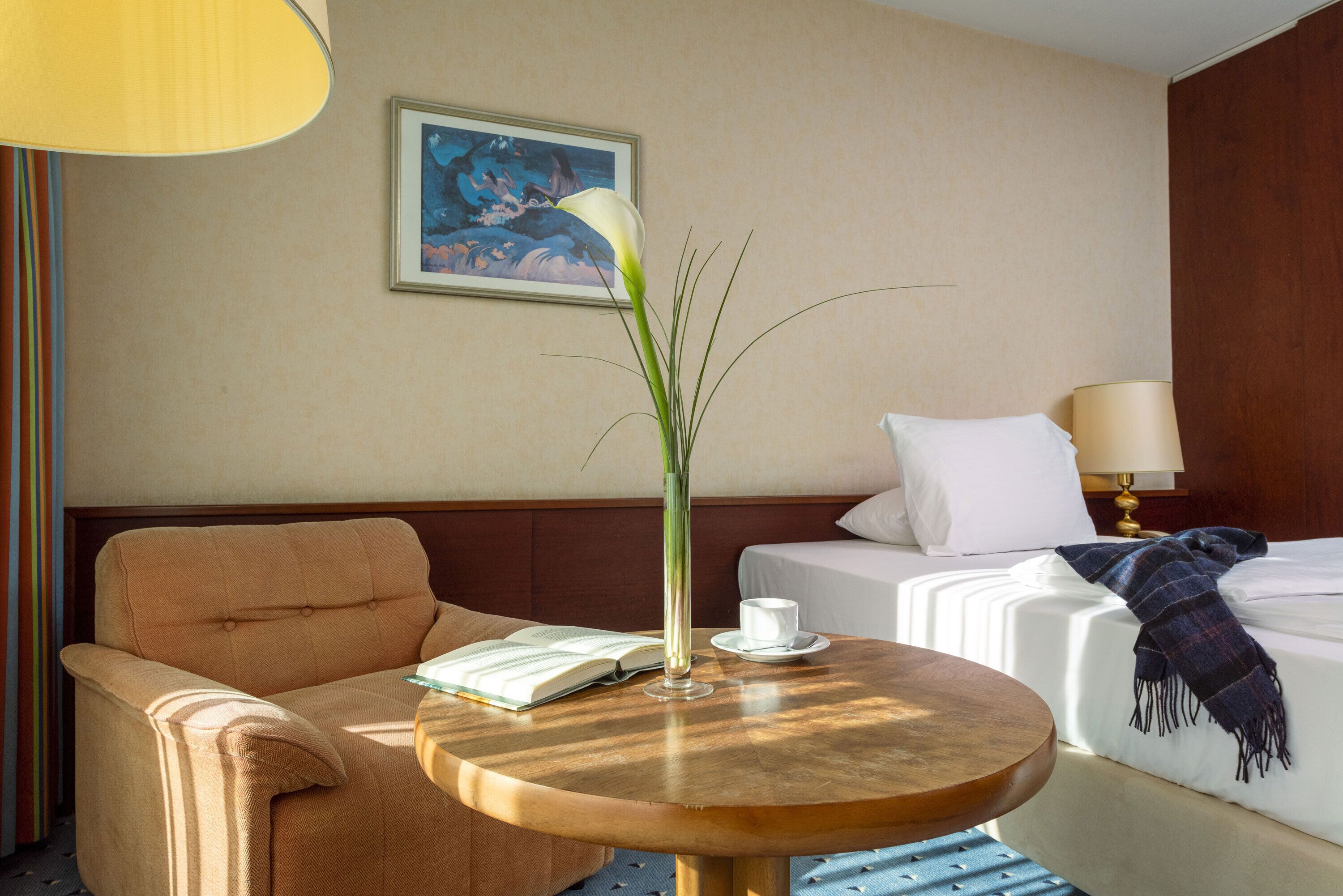 Ein Hotelzimmer mit einer Couch, einem runden Holztisch mit einem Buch und einer Vase, einem Bett mit weißer Decke und einem Bild an der Wand.