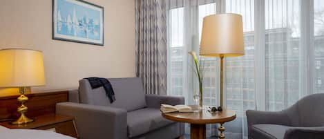 A hotel room with a bed, two armchairs, a sofa, a small round table with a lamp, a framed picture on the wall, and a window with sheer curtains.