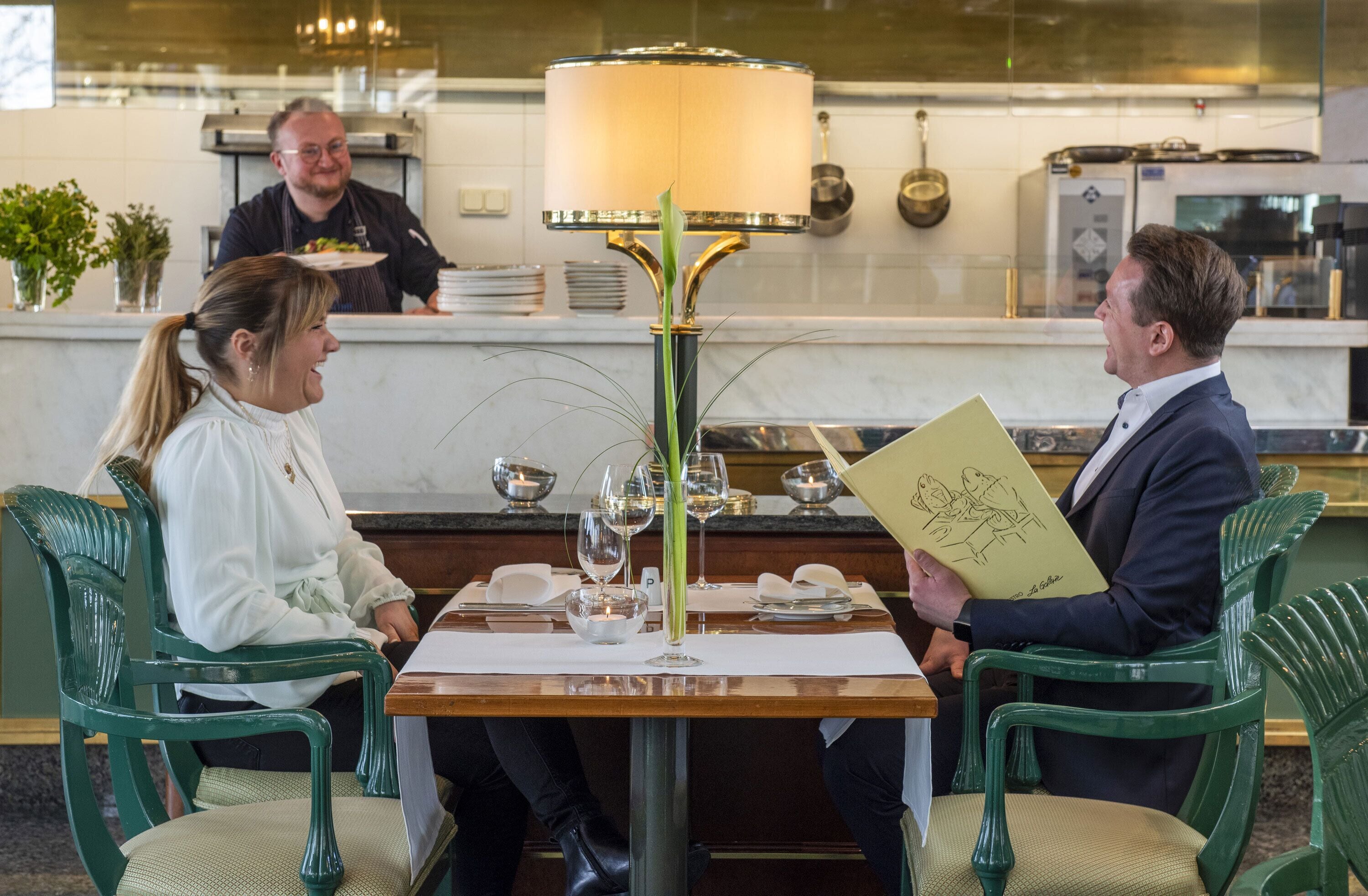 A dining table with a lamp, a menu, and a vase with green leaves.