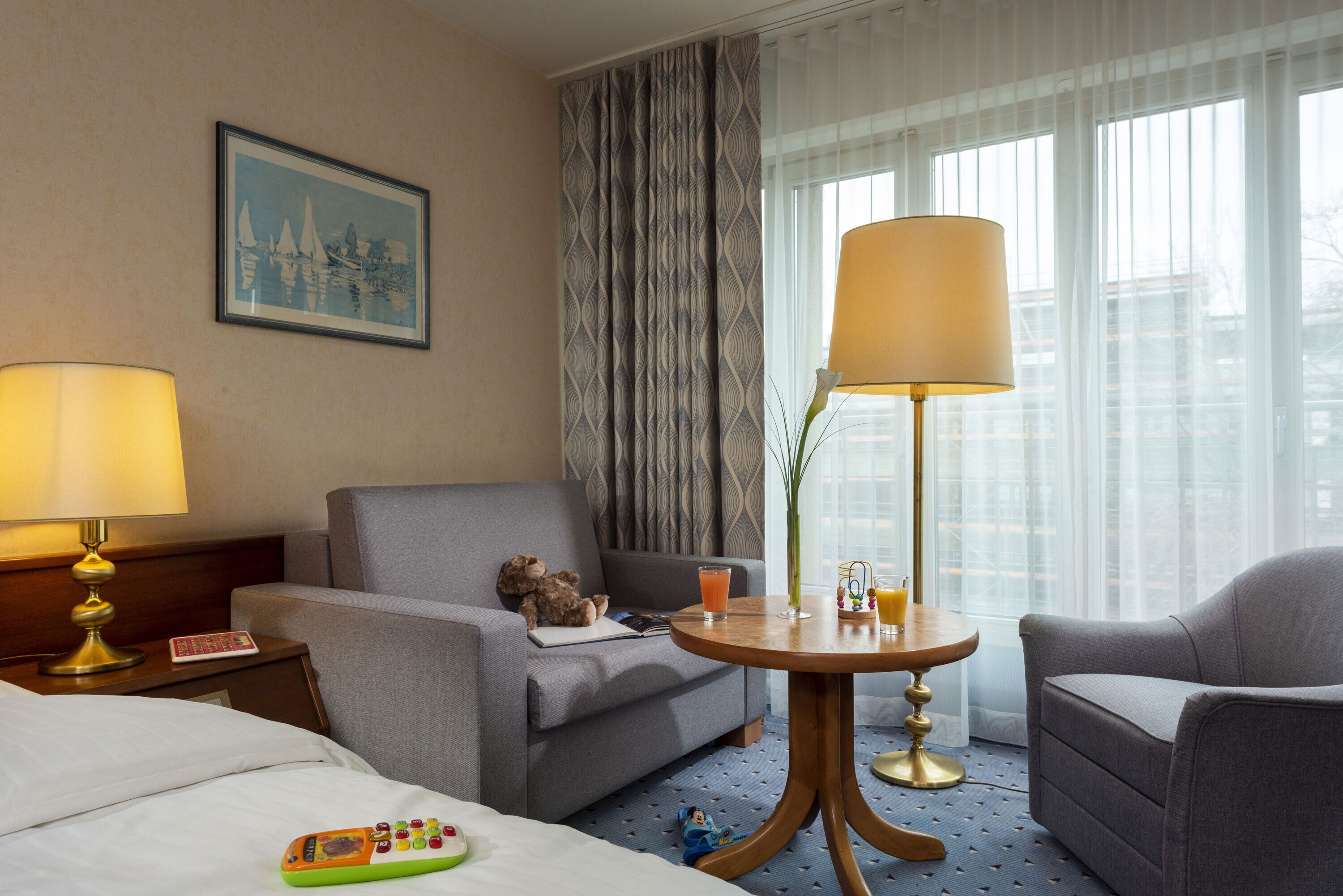 A hotel room with a bed, a sofa, a small table with a lamp, a chair, and a window with curtains.