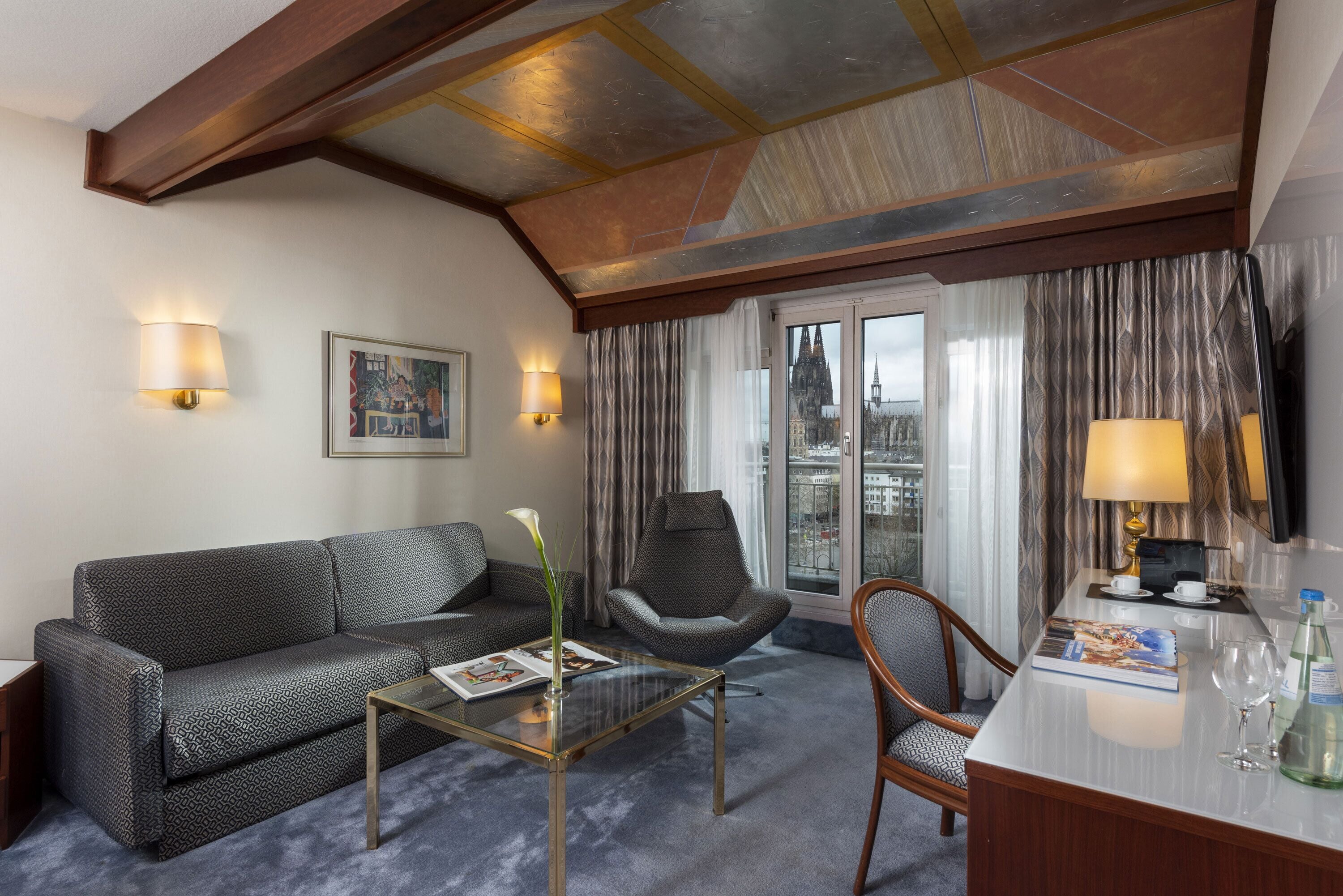 Habitación de hotel con sofá, sillón, mesa de centro de vidrio y un área de comedor con mesa y sillas.