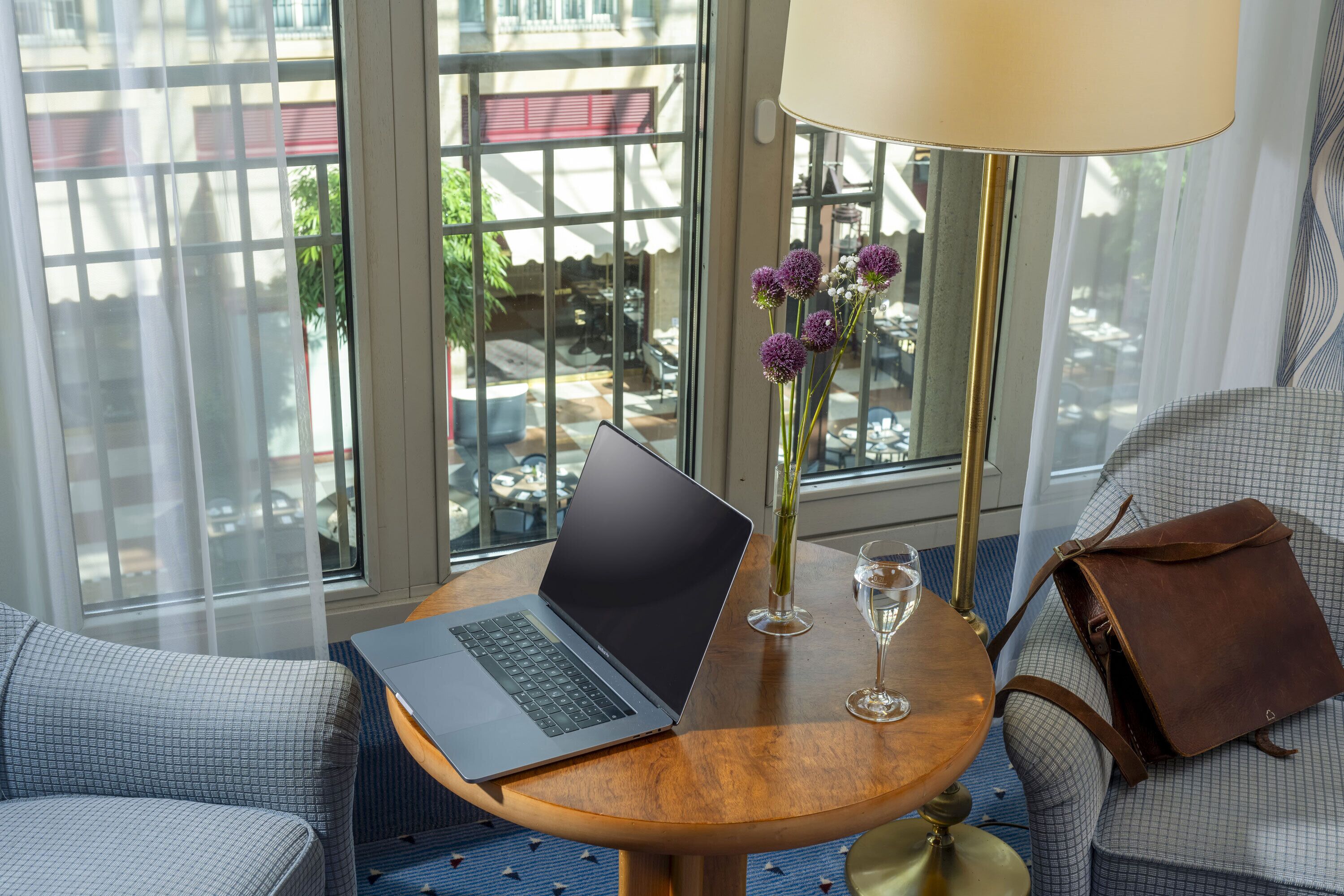 A laptop on a wooden table, a vase with purple flowers, a wine glass, and a brown leather bag on a checkered sofa.