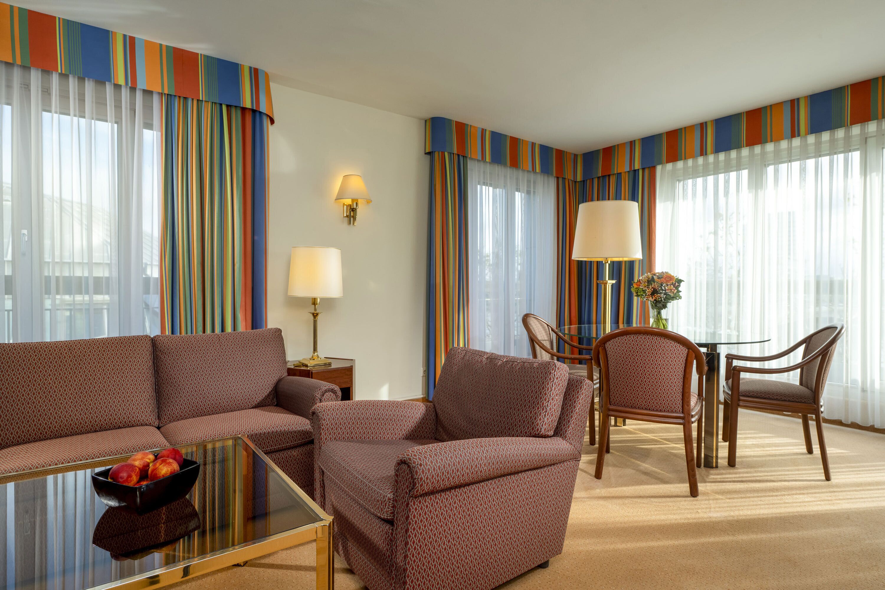A hotel room with a brown patterned sofa set, a glass coffee table with fruit, a dining table with chairs, and striped curtains.