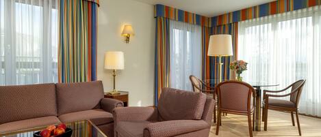 A hotel room with a brown patterned sofa set, a glass coffee table with fruit, a dining table with chairs, and striped curtains.