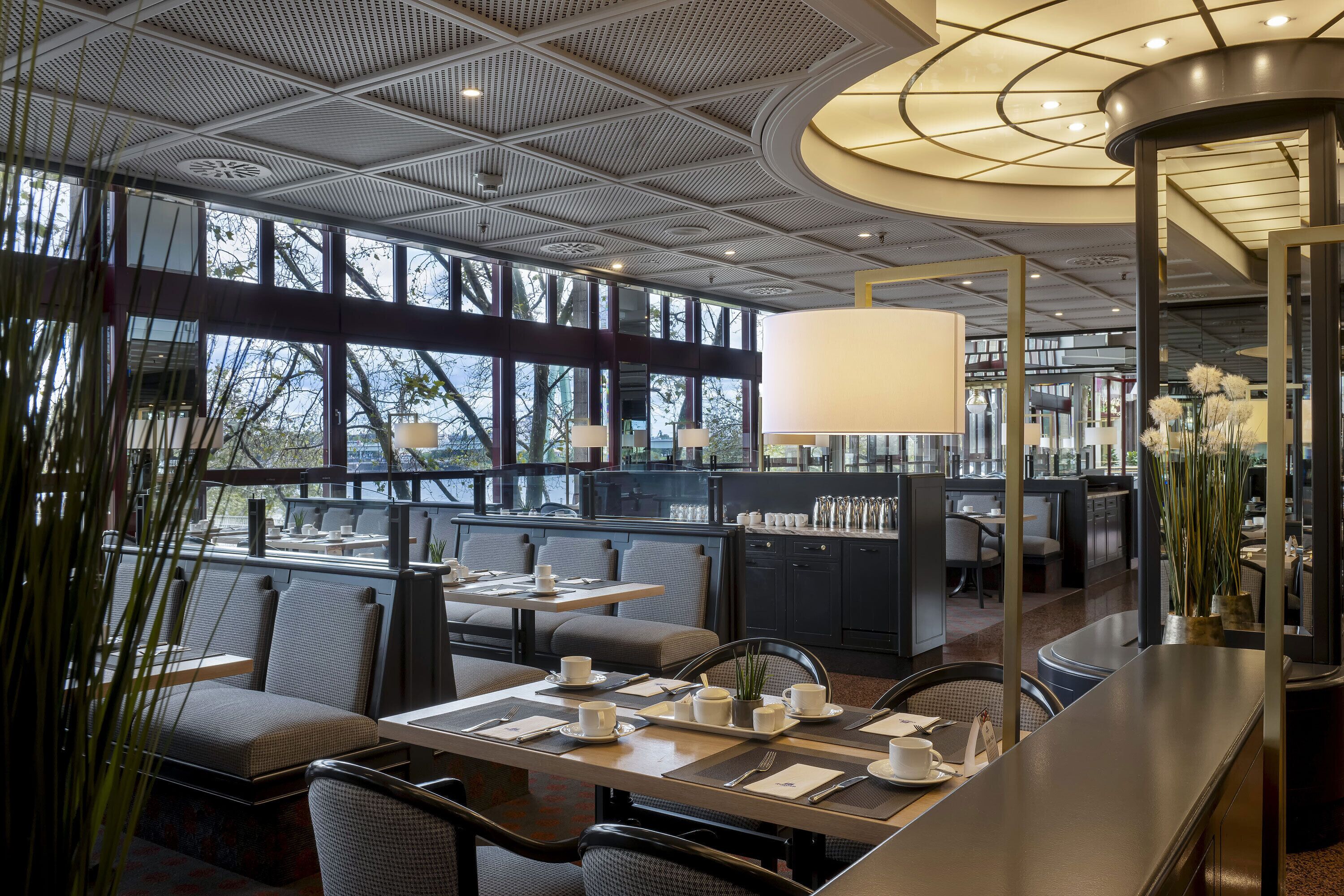 A modern restaurant with a high ceiling, large windows, and a central dining area with tables and chairs.