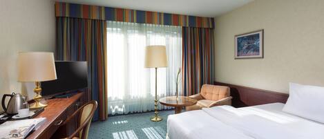 Habitación de hotel con cama, escritorio con lámpara, silla, televisor y ventana con cortinas.