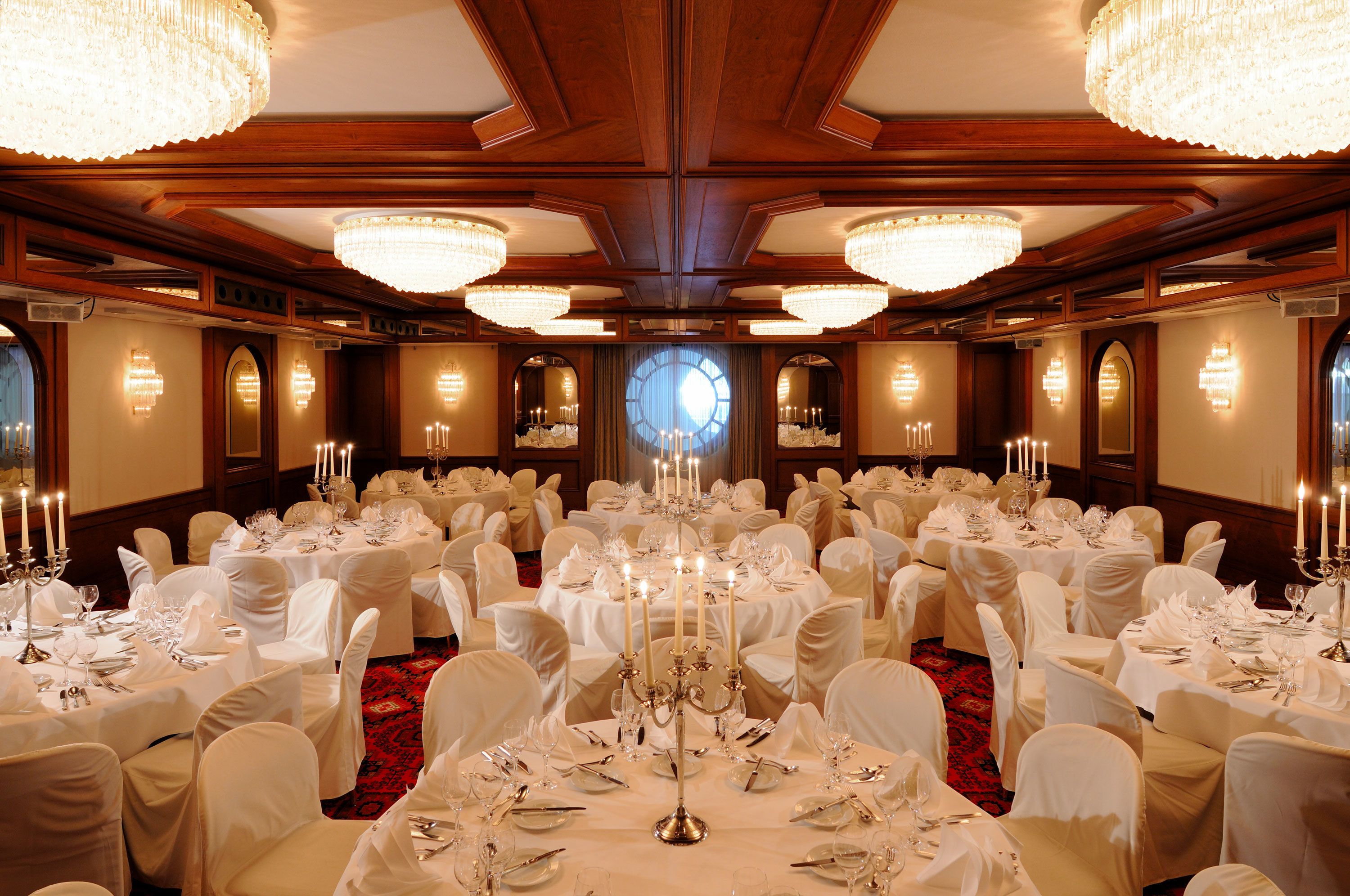 A banquet hall with round tables set for a formal event, featuring chandeliers and candle holders.