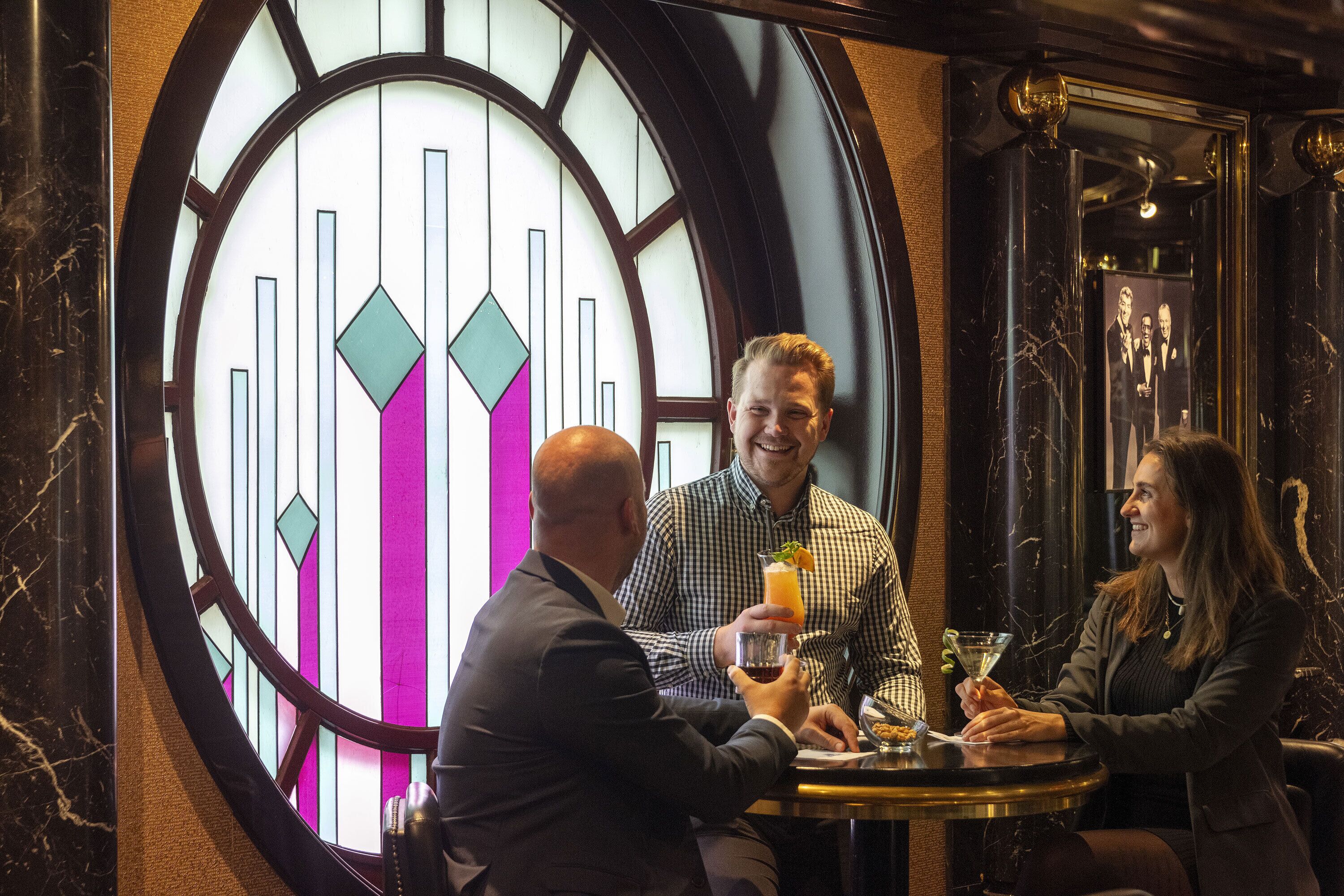 A man and a woman sitting at a table with drinks, in front of a large circular stained glass window with geometric patterns.