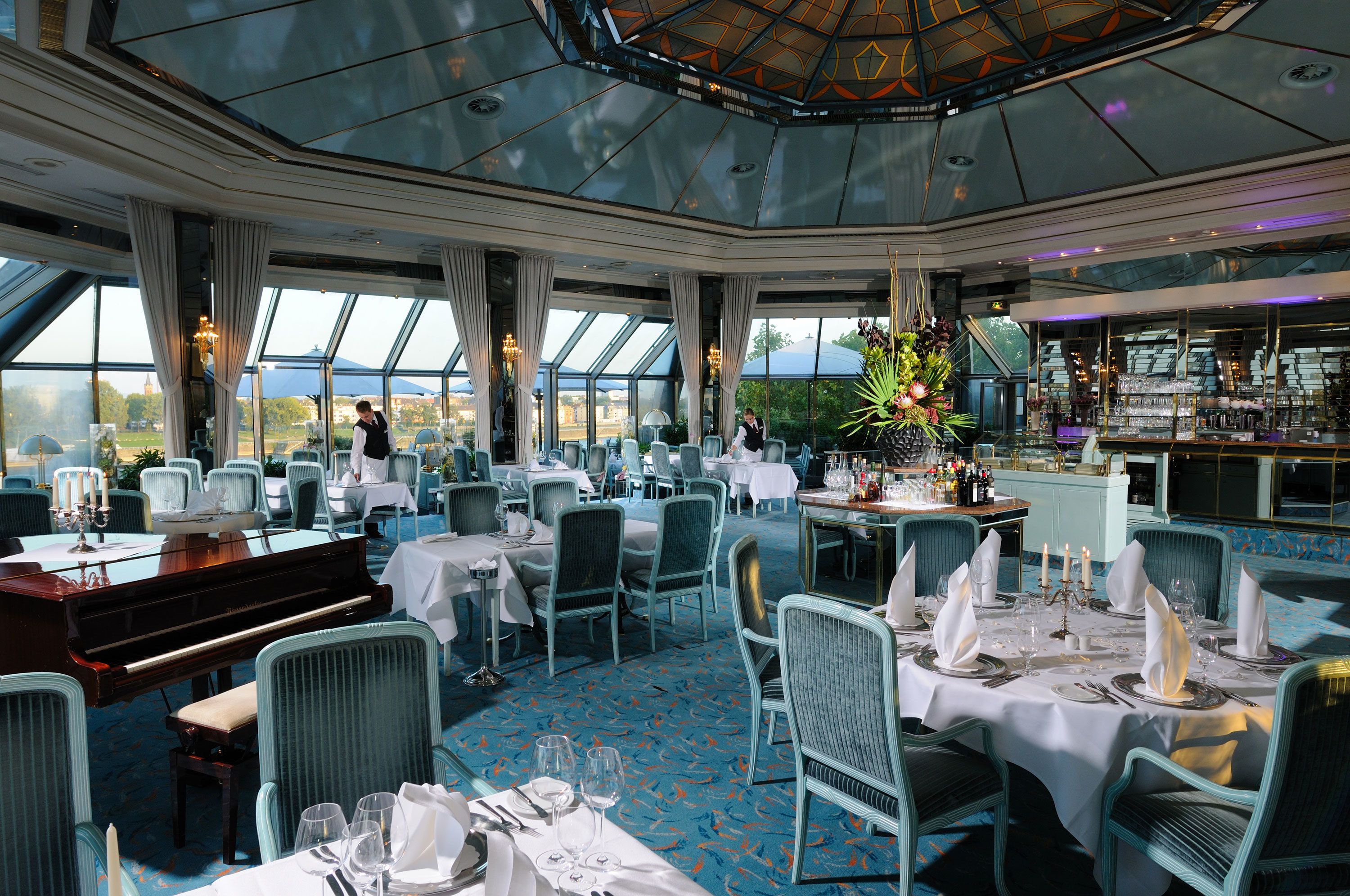 A restaurant with tables set for dining, a piano, and a bar area.
