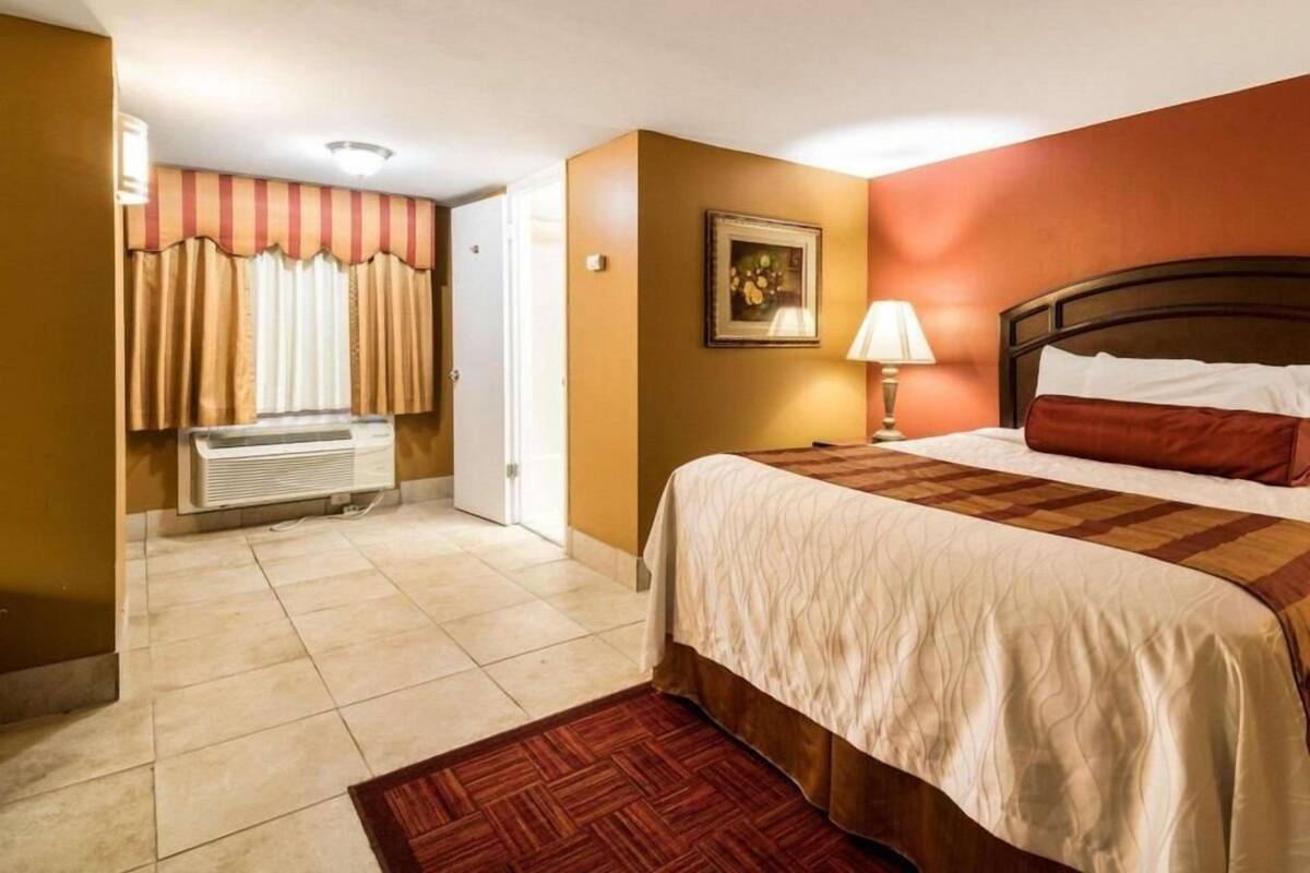 Ett hotellrum med en säng, ett nattduksbord, en lampa, en tavla på väggen och en dörr som leder till ett annat rum.