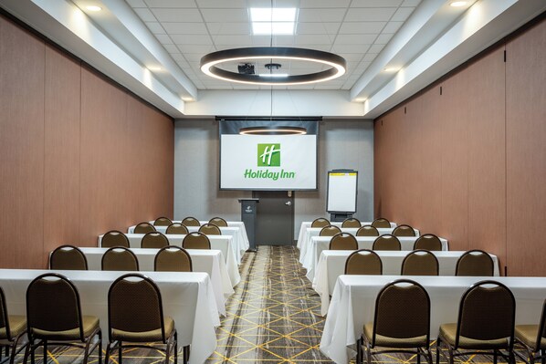 A conference room with rows of tables and chairs, a podium, and a screen displaying the Holiday Inn logo. - Holiday Inn Blytheville by IHG (Blytheville)