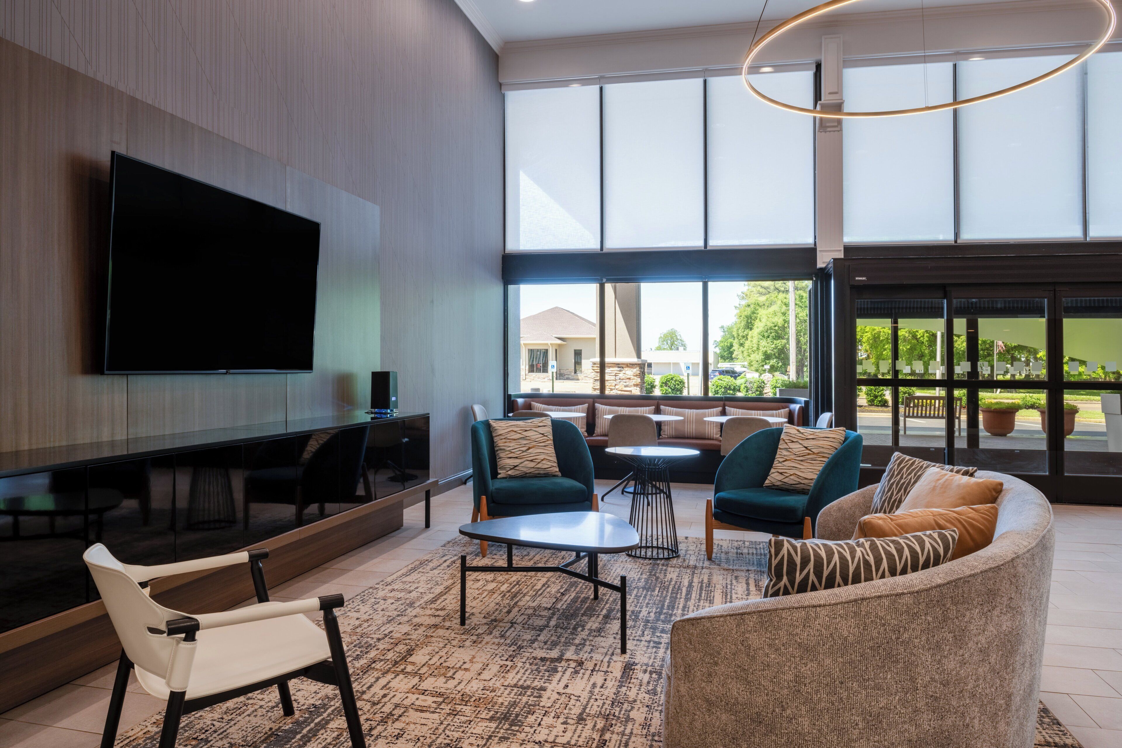 A modern living room with a flat-screen TV, comfortable seating, and large windows offering a view of the outdoors.
