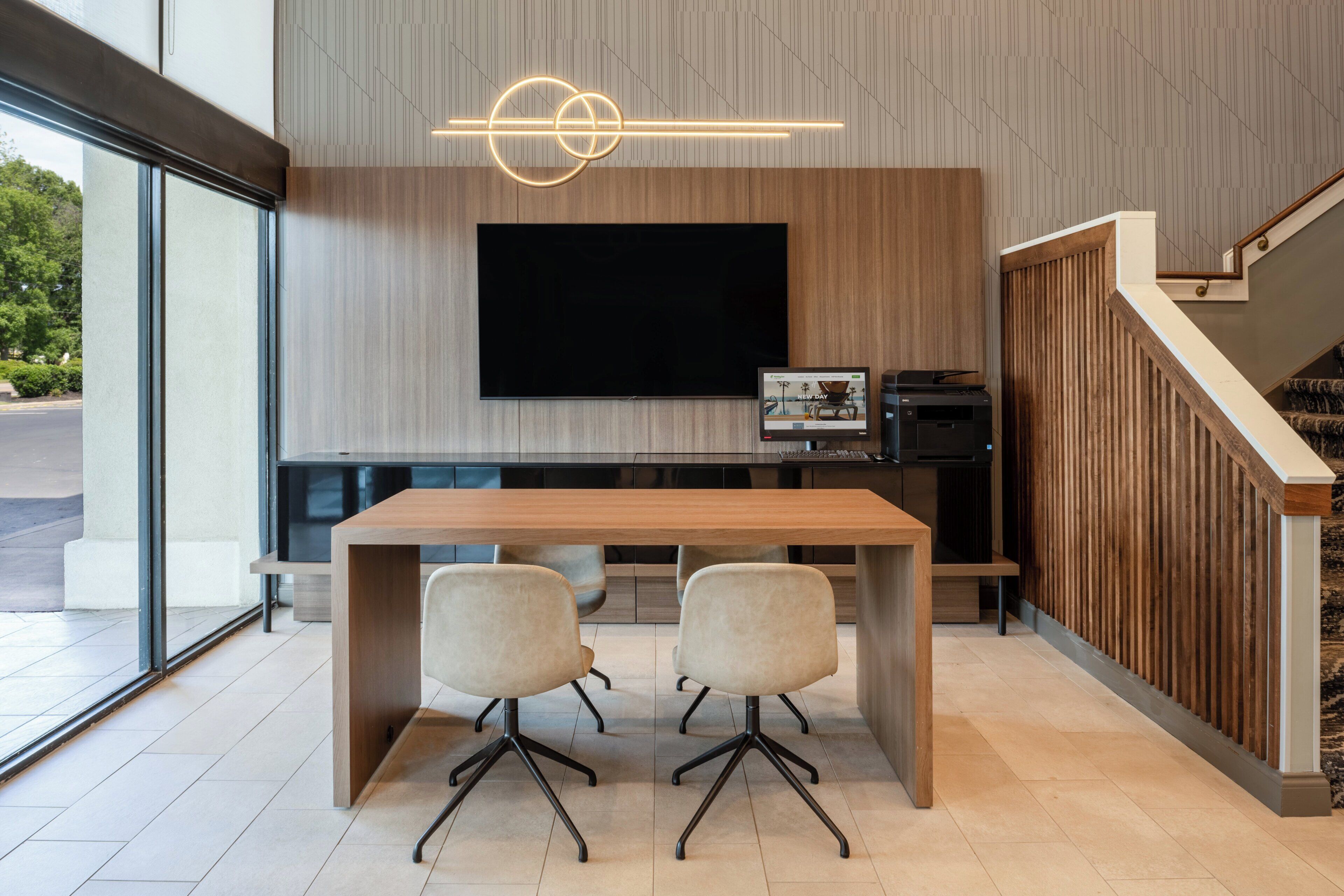 Un espacio de oficina moderno con un televisor de pantalla plana grande montado en una pared, una mesa de madera con dos sillas y una escalera con barandillas de madera.
