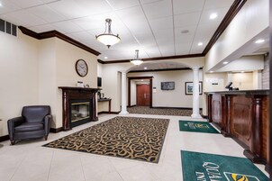 Un vestíbulo de hotel con una alfombra grande y estampada, una chimenea, un reloj en la pared y un mostrador de recepción.