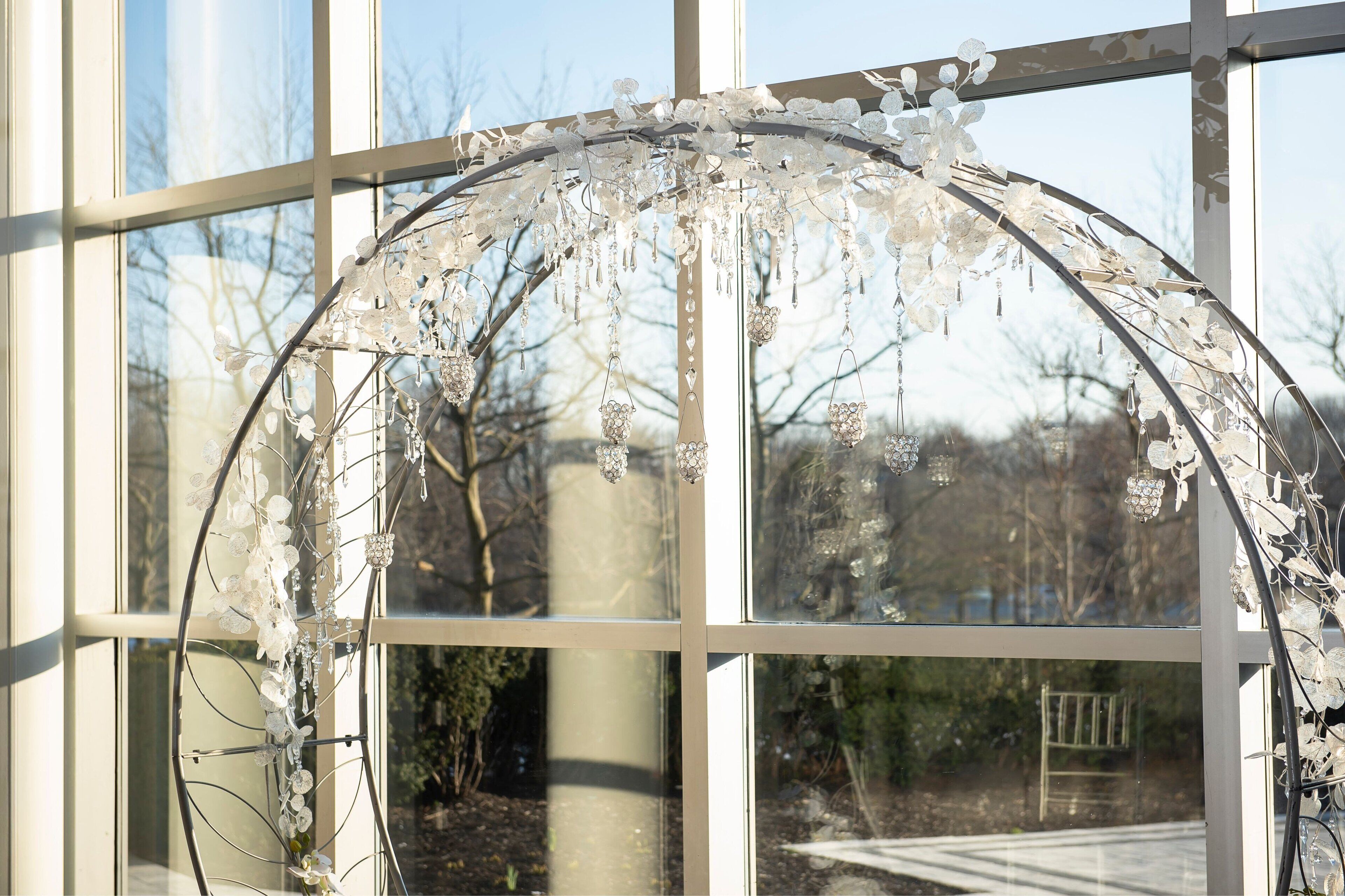Una estructura de vidrio decorativa con elementos colgantes, frente a una ventana que ofrece vistas a árboles y un sendero.