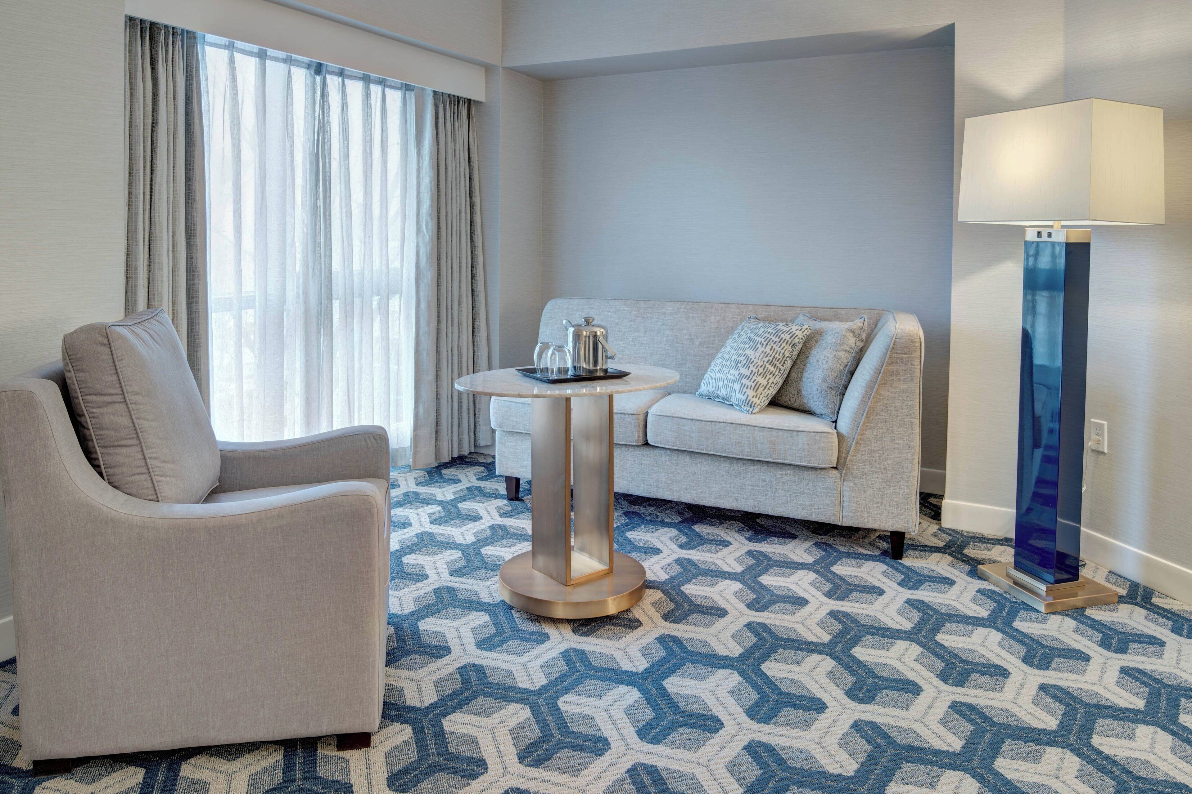 A hotel room with a sofa, armchair, round table with a tray, and a floor lamp.