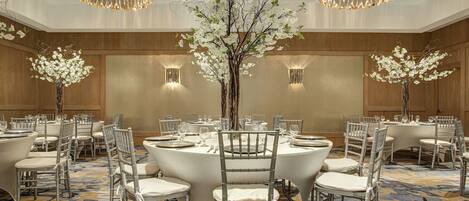 A banquet hall with round tables, white chairs, and floral centerpieces.