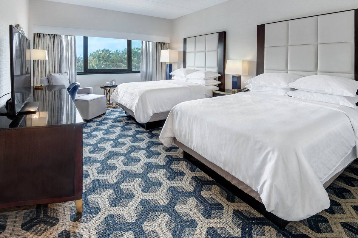 A hotel room with two beds, a desk, a chair, and a large window with curtains.