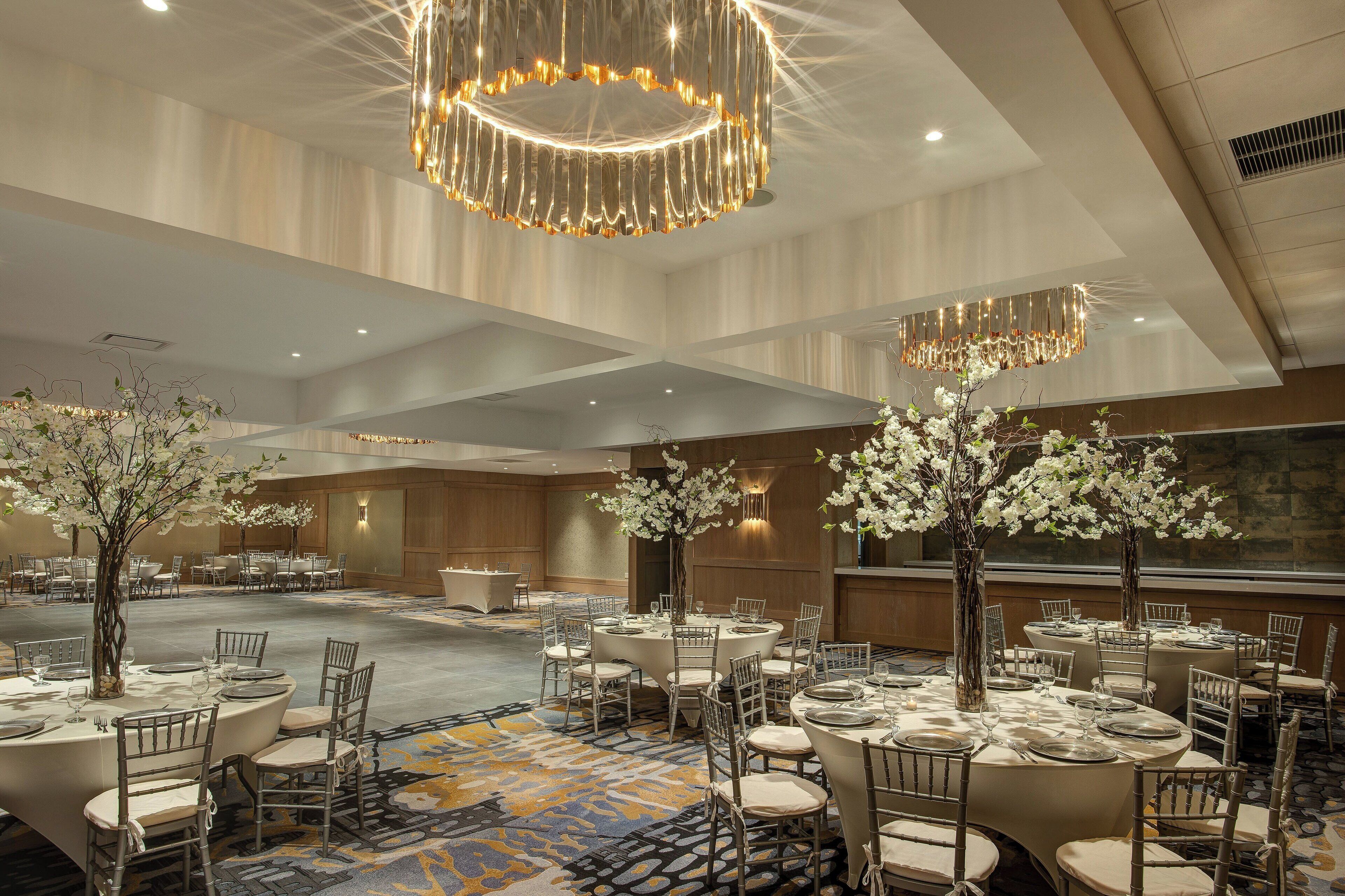 A spacious banquet hall with round tables, elegant chandeliers, and floral centerpieces.