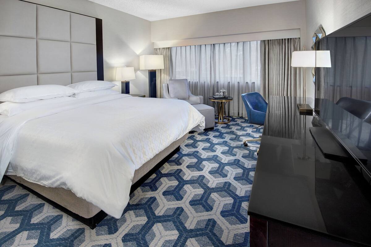 A hotel room with a large bed, a desk, a chair, a TV, and a window with curtains.