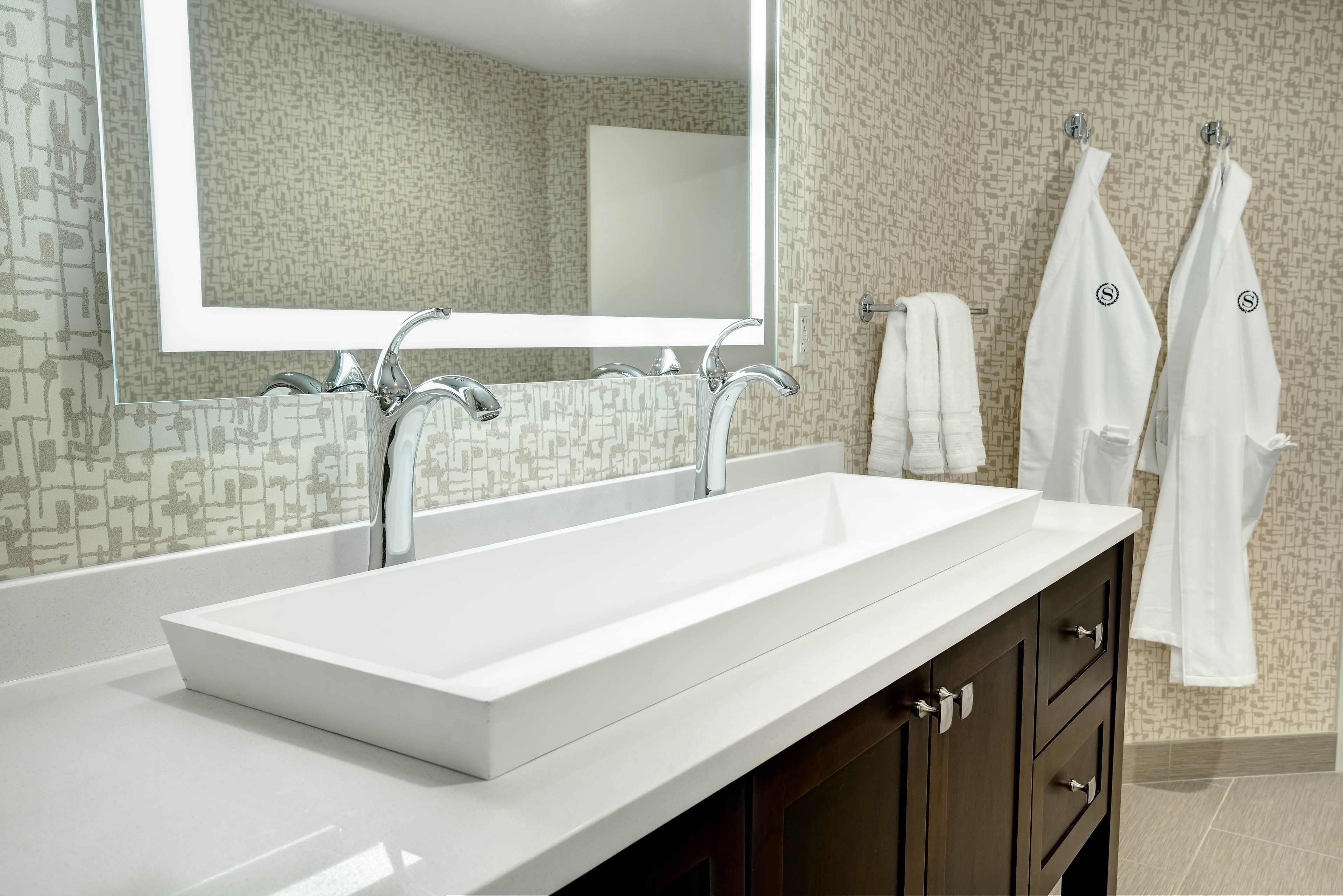 A modern bathroom with a large white sink, two faucets, a mirror, and a towel rack with two white towels.