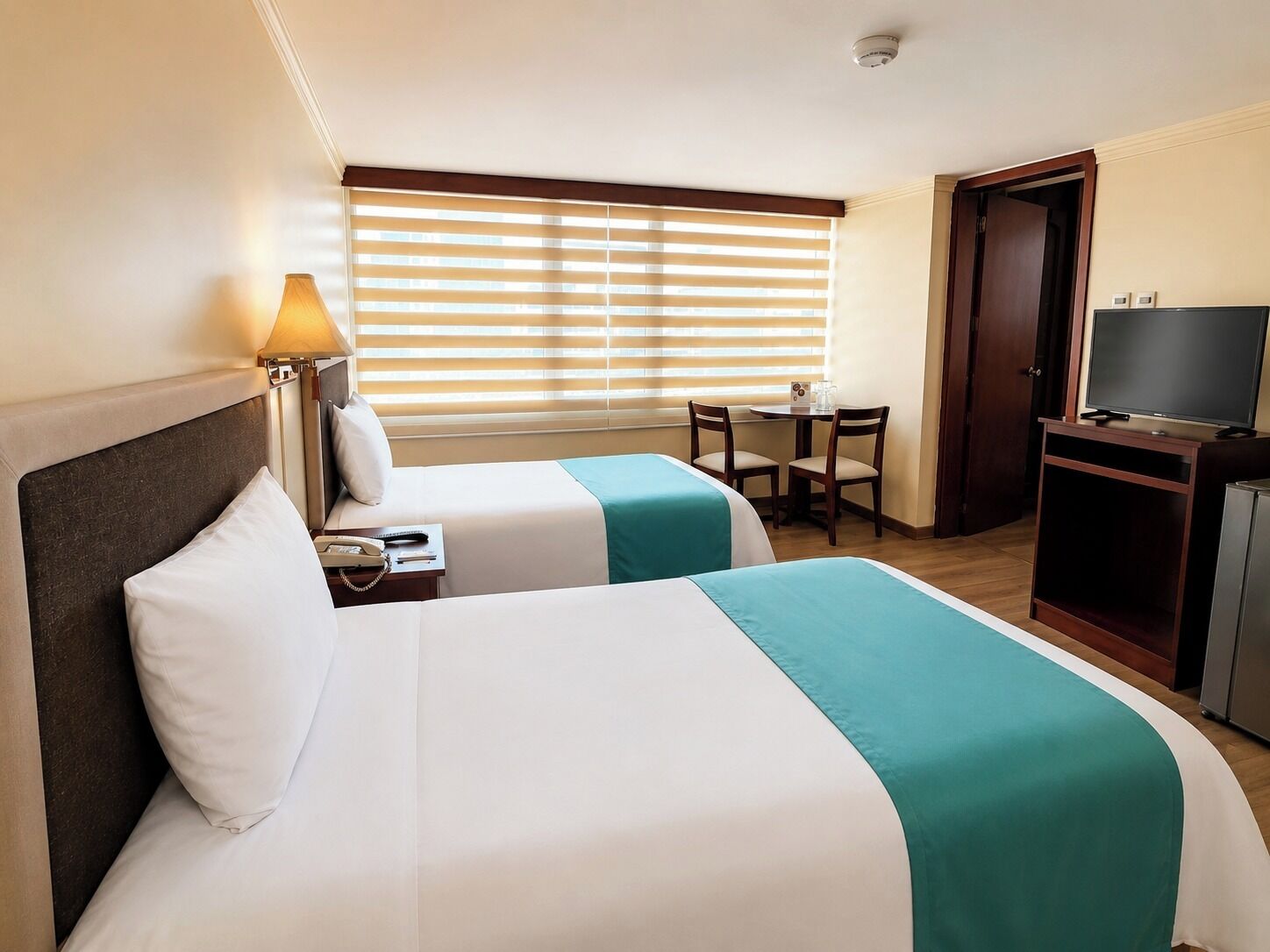 A hotel room with two beds, a desk with chairs, a television, and a window with blinds.
