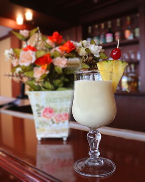 A cocktail in a glass with a pineapple garnish and a cherry on top, placed on a bar counter with a floral arrangement in the background. - Hotel República (Quito)