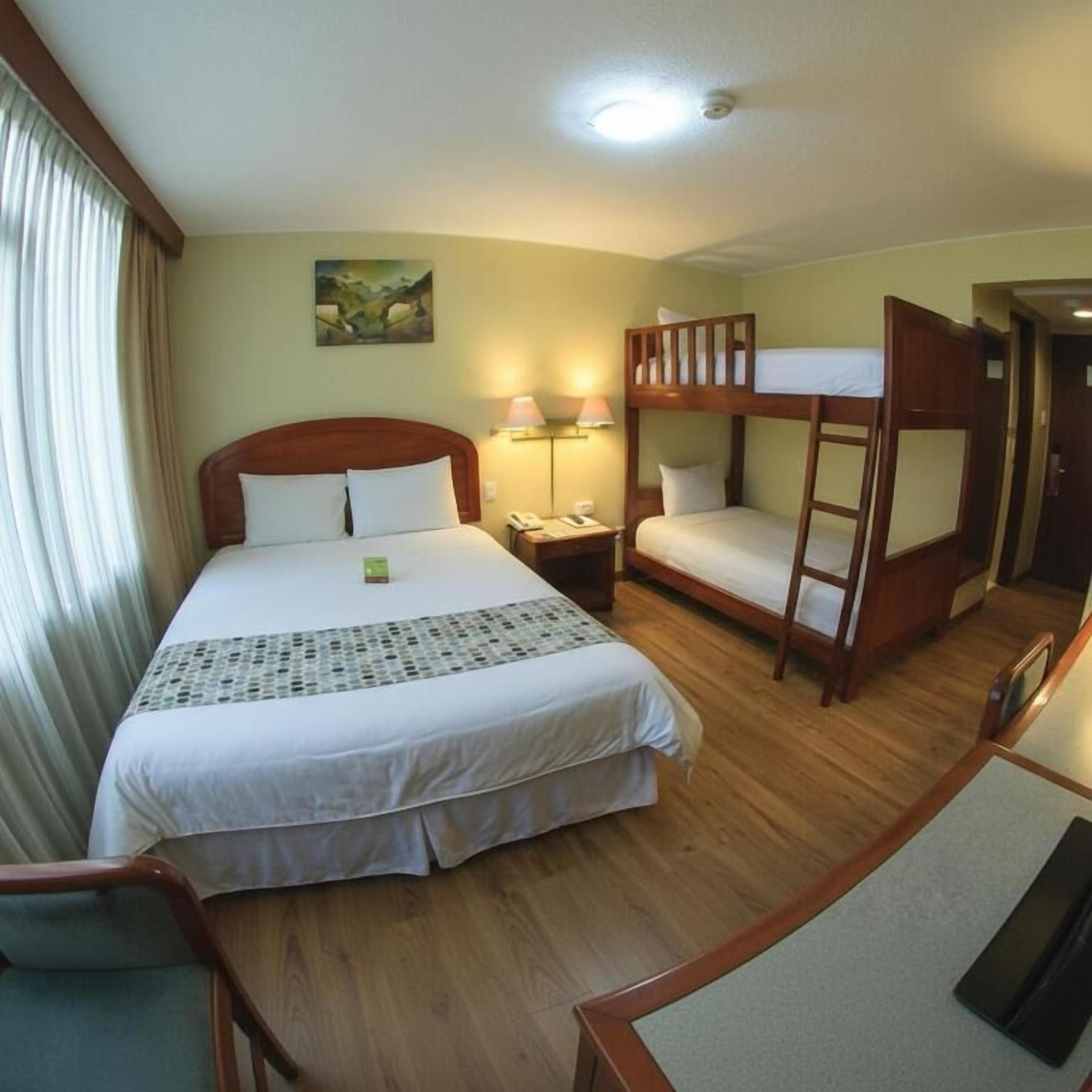 Una camera d'albergo con un letto singolo, un letto a castello, una scrivania con una sedia, una lampada e un quadro appeso al muro.