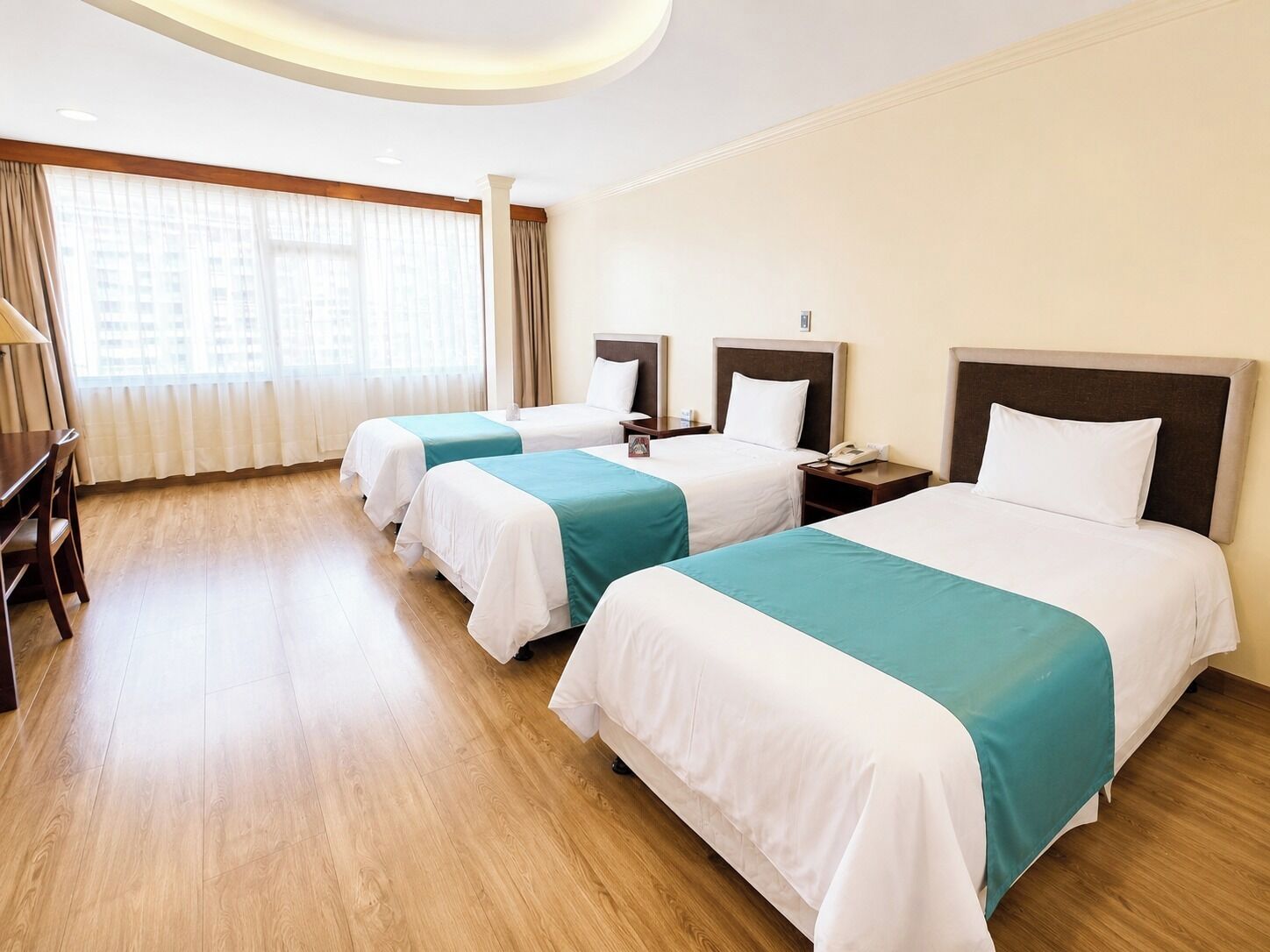 A hotel room with three beds, a desk, and a large window with curtains.