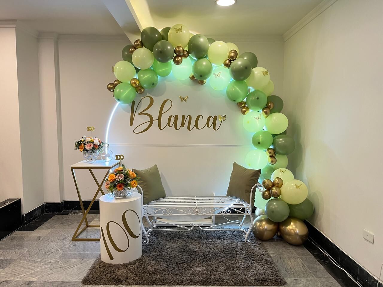 Una habitación con un arco decorativo de globos, una cama blanca con estructura metálica, una mesita con un florero y un objeto cilíndrico con el número 100.