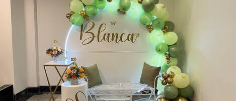 Una habitación con un arco decorativo de globos, una cama blanca con estructura metálica, una mesita con un florero y un objeto cilíndrico con el número 100.