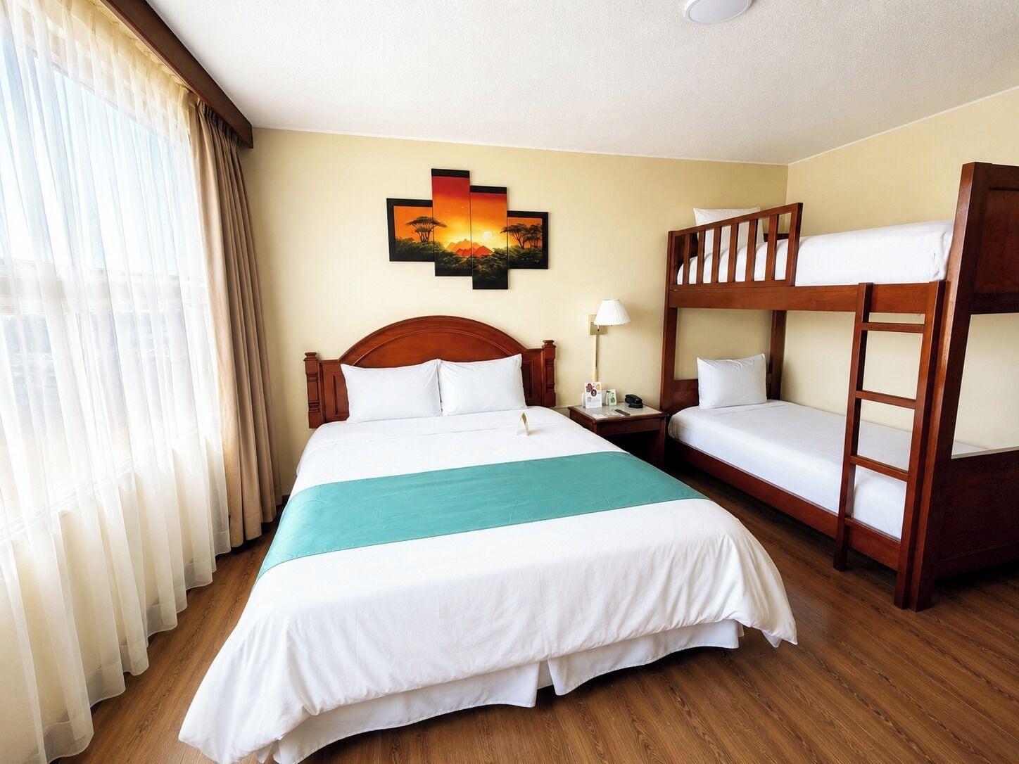 A hotel room with a large bed, a bunk bed, a painting on the wall, and a window with curtains.