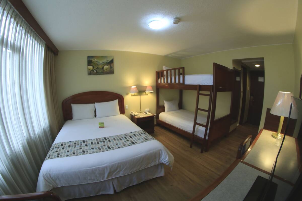 Une chambre d’hôtel avec un grand lit, un lit superposé plus petit, un bureau avec une lampe et un tableau au mur.