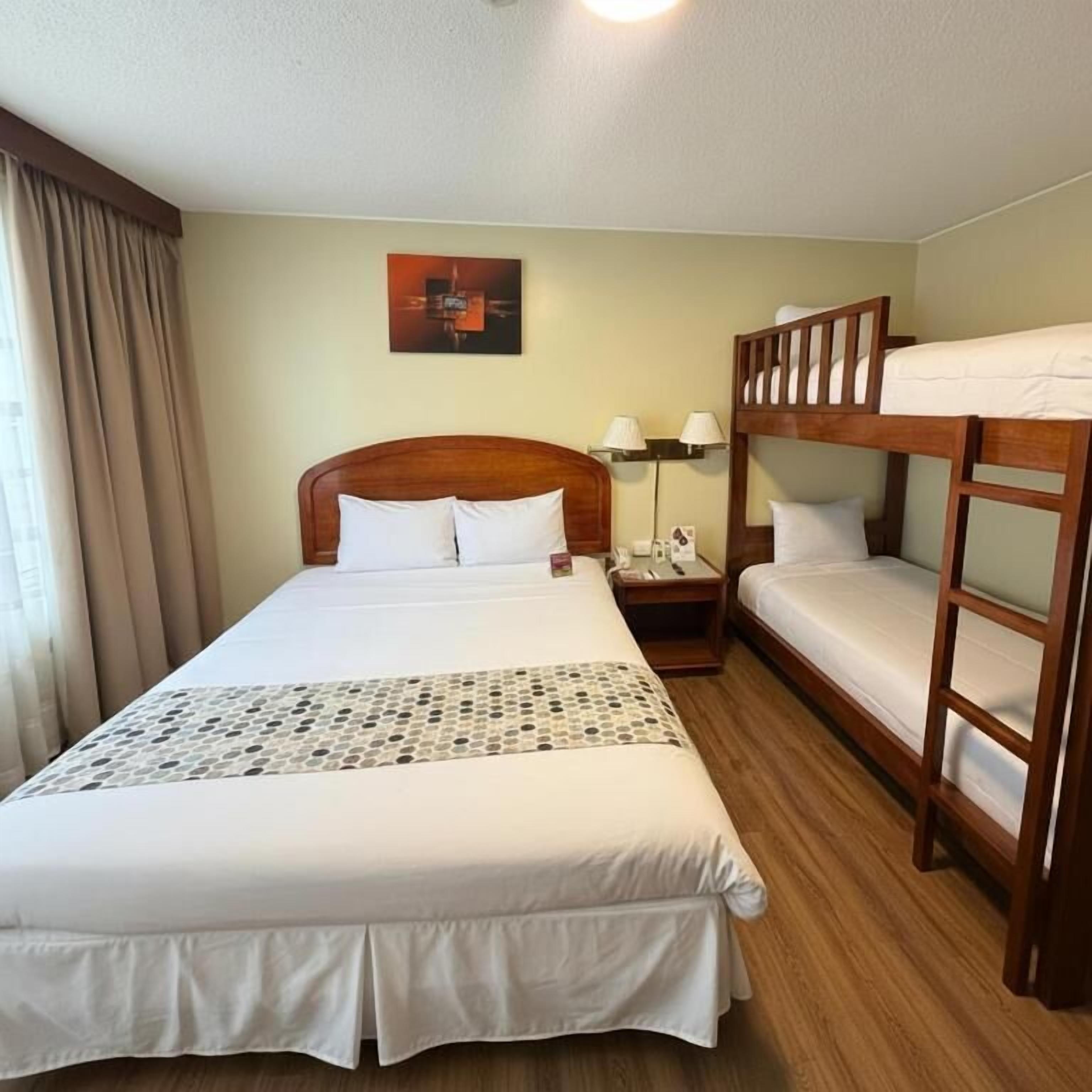 A hotel room with a large bed, a bunk bed, a bedside table with a lamp, and a wall-mounted painting.