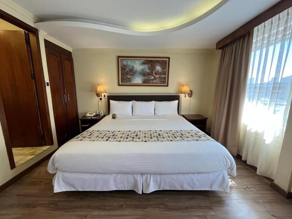 A hotel room with a large bed, wooden wardrobe, a painting on the wall, and a window with curtains.