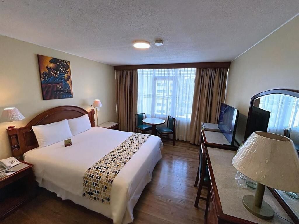 Ein Hotelzimmer mit einem großen Bett, einem Schreibtisch mit Stuhl, einem Fernseher und einem Fenster mit Vorhängen.