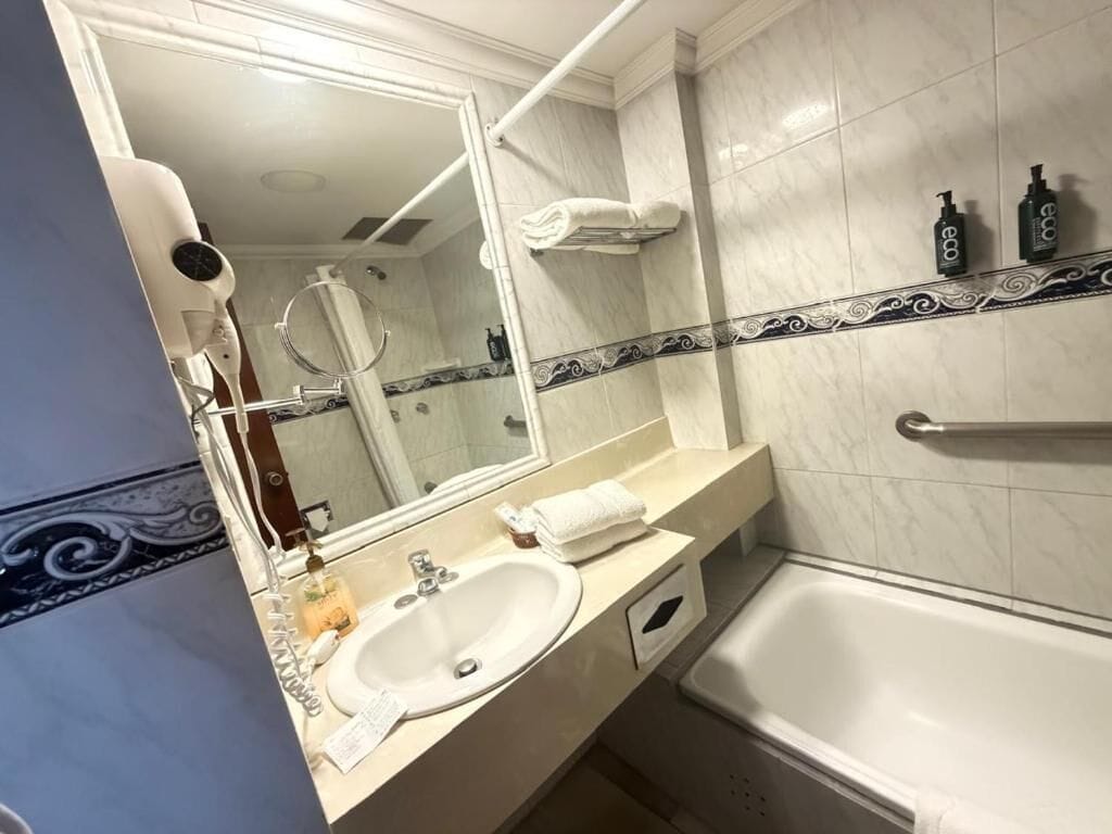 A bathroom with a white sink, mirror, and bathtub. There are two bottles on the wall, a towel rack, and a towel on the counter.