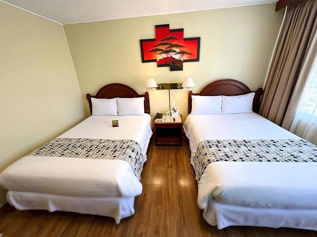 A hotel room with two single beds, a bedside table with a lamp, a wall-mounted artwork, and a window with curtains.