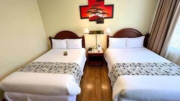 A hotel room with two single beds, a bedside table with a lamp, a wall-mounted artwork, and a window with curtains.