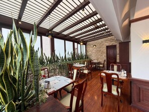 Un comedor interior con mesas y sillas de madera, pared de piedra y ventanas amplias.